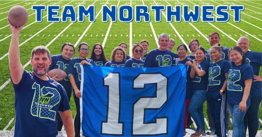 A vibrant image displaying a group of enthusiastic individuals wearing blue t-shirts with "NW Northwest" logo on a football field. The word "TEAM NORTHWEST" is prominently displayed at the top. In the foreground, a man holding a football high in his left hand stands out. Beside him, the group holds up a large blue flag featuring the number "12". Everyone in the photo looks happy and spirited, radiating team spirit.
